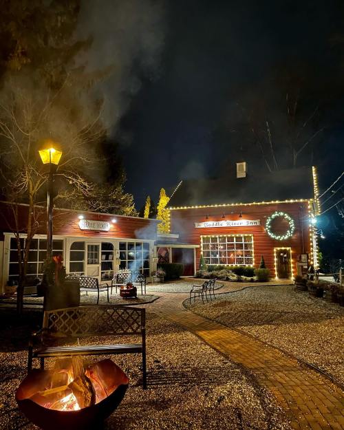 A cozy inn at night, adorned with lights, surrounded by a fire pit and benches, creating a warm, inviting atmosphere.