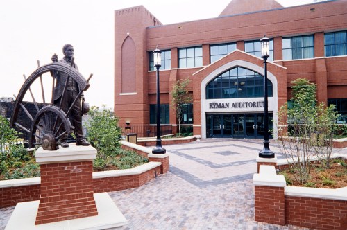 Statue of a man with a wheel in front of the Ryman Auditorium, a brick building with large windows and landscaped pathways.