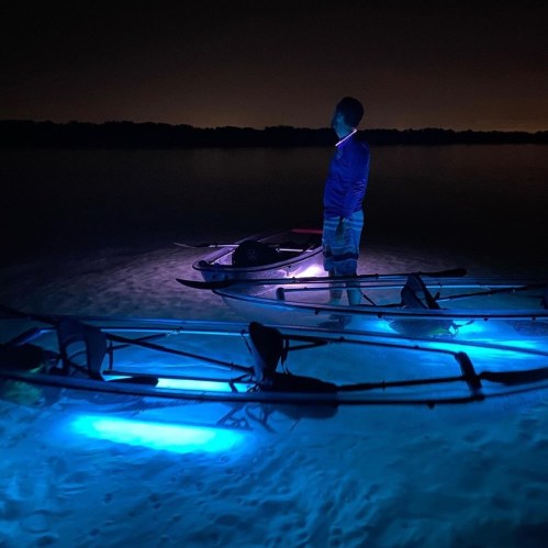 A person stands on a beach at night near illuminated kayaks, with a calm water backdrop and a glowing horizon.