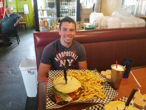 A smiling man sits at a table with a large burger, fries, and drinks in a casual restaurant setting.