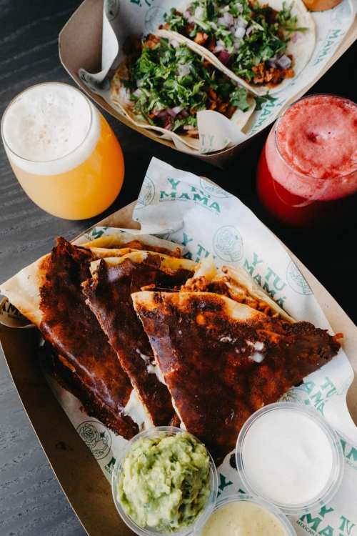 A vibrant spread of tacos, quesadillas, and drinks, featuring guacamole and sauces on a wooden table.