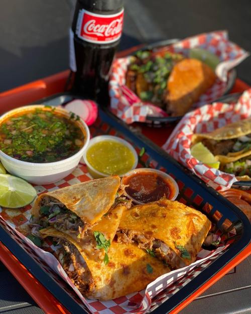 A vibrant tray with tacos, a bowl of soup, sauces, lime, and a bottle of Coca-Cola, all served on a checkered paper.