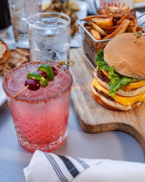 A vibrant pink cocktail with garnishes beside a cheeseburger and fries on a wooden board.