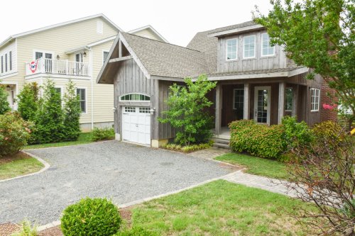 A charming wooden house with a gravel driveway, surrounded by greenery and another house nearby.