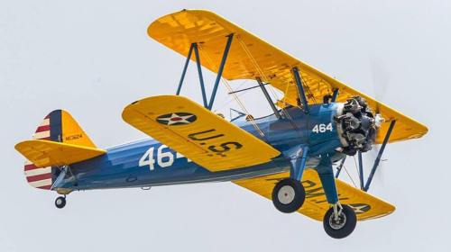A vintage biplane with a blue body and yellow wings flying against a light sky. The plane has U.S. markings and the number 464.