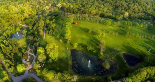 Aerial view of a lush green park with ponds, fountains, and winding paths surrounded by trees.
