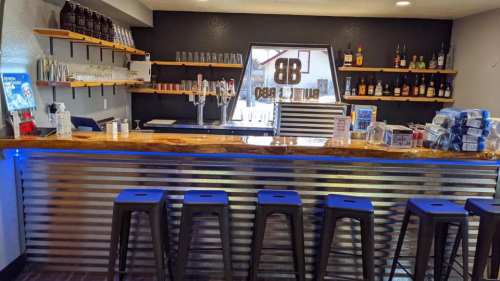 A modern bar with a wooden countertop, metal accents, shelves of liquor, and black stools in a stylish setting.