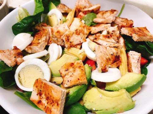 A colorful salad with grilled chicken, avocado, boiled eggs, baby corn, and mixed greens in a white bowl.