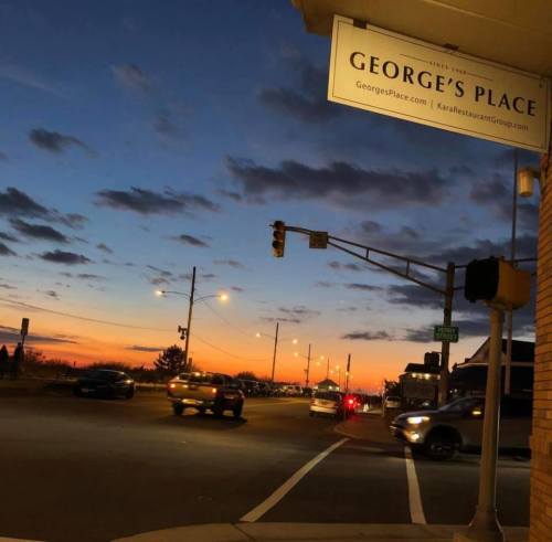 A street corner at dusk with a colorful sunset, traffic lights, and cars passing by near George's Place restaurant.