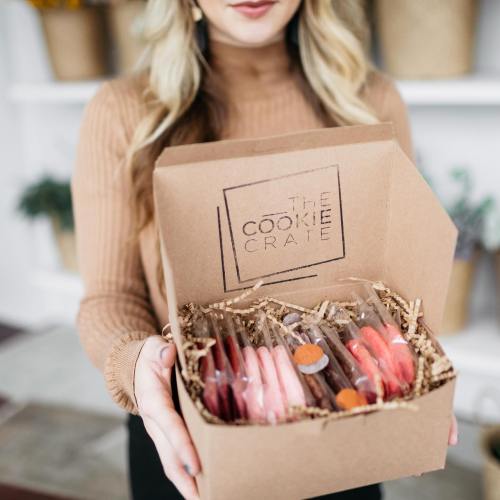 A person holds a box labeled "The Cookie Crate" filled with colorful cookies and decorative packaging.