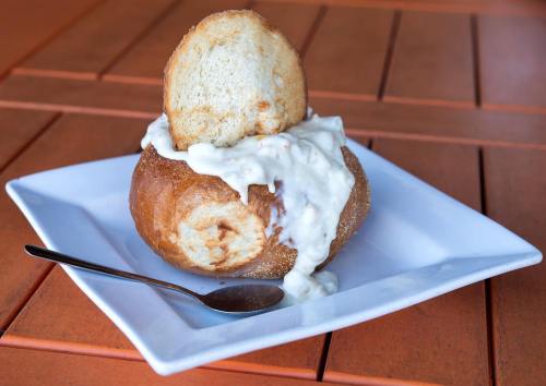 A bread bowl filled with creamy soup, topped with a slice of toasted bread, on a white plate with a spoon.