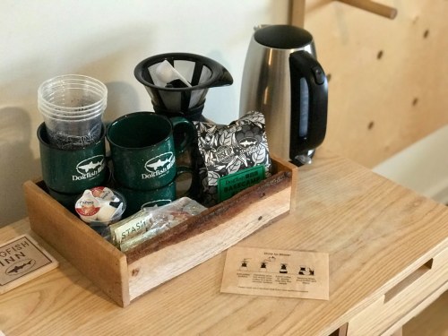 A wooden tray with mugs, tea bags, creamers, and a kettle on a table, with a guide for brewing coffee nearby.
