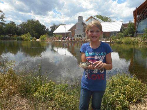A smiling girl holds a fish by a pond, with a building and people in the background under a cloudy sky.
