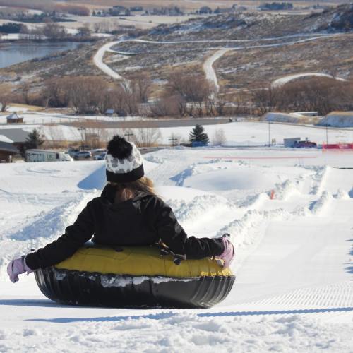 A person in a black and white hat sits in a yellow snow tube, ready to slide down a snowy hill.