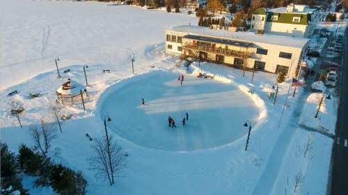 Aerial view of a snowy outdoor ice rink with skaters, surrounded by snow-covered ground and a building nearby.