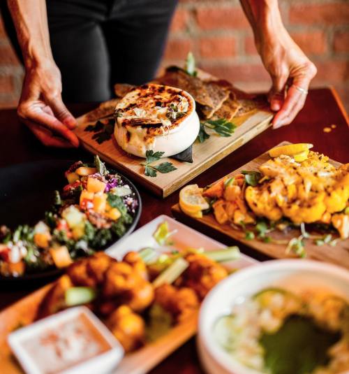 A person serves a variety of colorful dishes, including grilled vegetables, salads, and a creamy cheese on a wooden board.