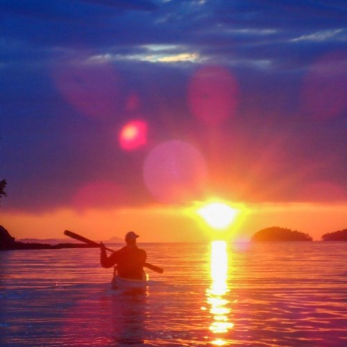 A kayaker paddles towards a vibrant sunset over calm waters, with colorful reflections and silhouettes of distant islands.