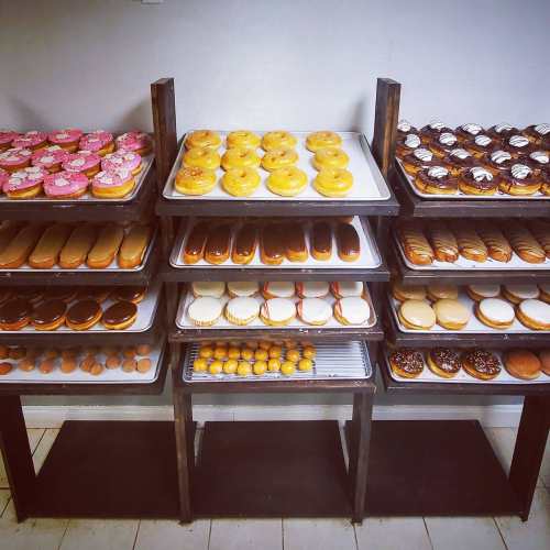 A display of various donuts on trays, featuring colorful toppings and different shapes, arranged on metal racks.