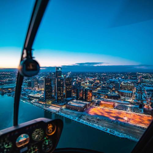 Aerial view of a city skyline at dusk, with lights reflecting on the water and a helicopter cockpit in the foreground.
