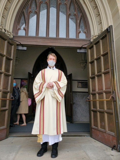 A priest in a white robe stands at the entrance of a church, wearing a mask, with people visible in the background.