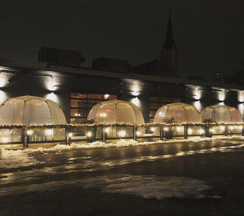 A row of illuminated outdoor dining igloos at night, with snow on the ground and a building in the background.