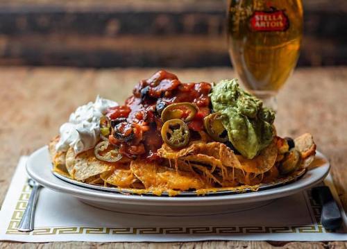 A plate of loaded nachos topped with cheese, jalapeños, guacamole, sour cream, and salsa, alongside a beer.