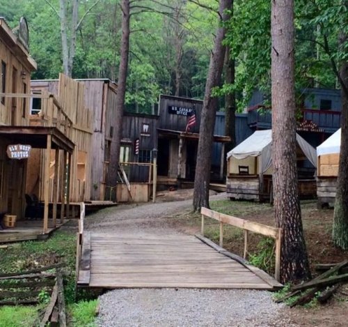 A rustic pathway leads through a wooded area with wooden buildings and tents, evoking a historical or western theme.