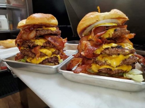 Two towering burgers stacked with multiple beef patties, cheese, bacon, and toppings on a metal tray.