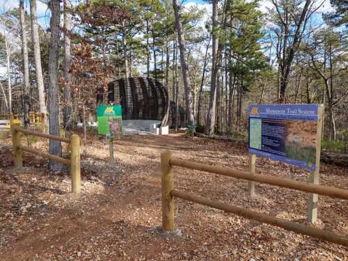 Entrance to a trail system with signs and a wooden structure, surrounded by trees and gravel path.