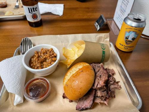 A tray with sliced brisket, a bun, baked beans, potato chips, and a beer can on a wooden table.