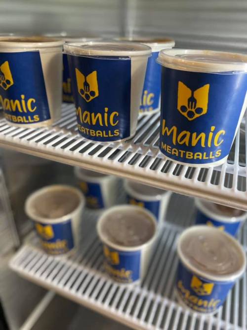 Cups of "Manic Meatballs" stacked in a refrigerator, showcasing a blue and yellow logo.