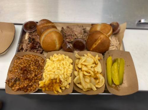 A tray of barbecue with pulled meat, buns, baked beans, macaroni and cheese, onion rings, and pickles.