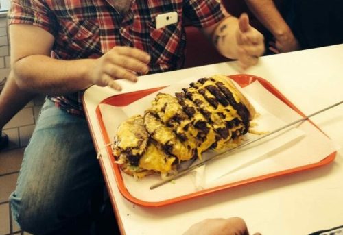 A man in sunglasses gestures excitedly at a large, cheesy meat-filled dish on a tray in a casual dining setting.