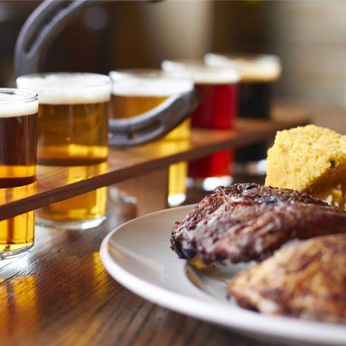 A wooden flight of beer glasses alongside a plate of grilled meat and cornbread on a table.