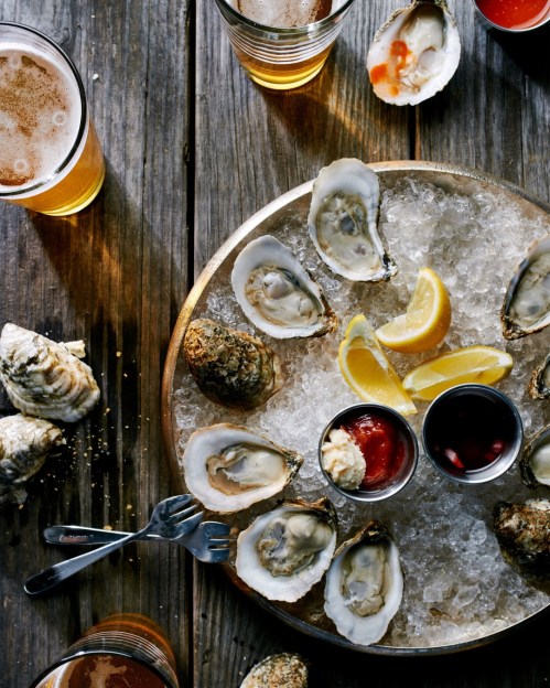 A platter of oysters on ice with lemon wedges and dipping sauces, surrounded by glasses of beer on a wooden table.