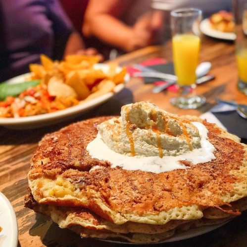 A stack of large pancakes topped with whipped cream and syrup, with plates of food and drinks in the background.