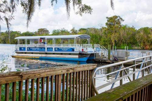 A boat docked at a riverside, surrounded by trees and greenery, with a wooden railing in the foreground.