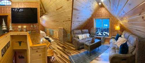 Cozy wooden cabin interior with a living area, plush sofas, a TV, and large windows showcasing a snowy landscape.