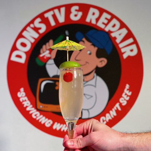 A hand holds a cocktail with a cherry and lime, featuring a small umbrella, in front of a colorful sign for Don's TV & Repair.