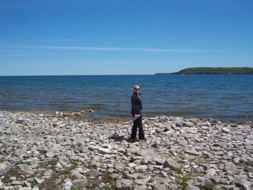 A person in a suit stands on rocky shore by a calm blue lake under a clear sky.