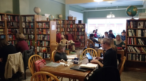 A cozy bookstore with people reading and working at tables surrounded by shelves of books.