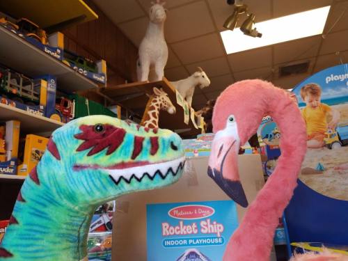 A colorful dinosaur plush toy and a pink flamingo plush toy face each other in a toy store filled with animal figures.