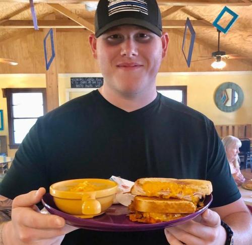 A smiling man holds a plate with grilled cheese sandwiches and a bowl of macaroni and cheese in a cozy dining setting.