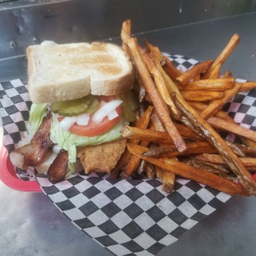 A sandwich with lettuce, tomato, pickles, and bacon, served with a side of crispy sweet potato fries in a checkered basket.