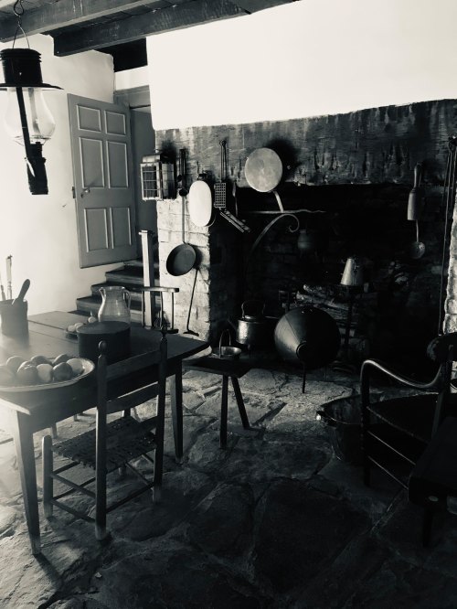 A rustic kitchen with a wooden table, chairs, and a large stone fireplace, featuring vintage cooking tools and a lantern.