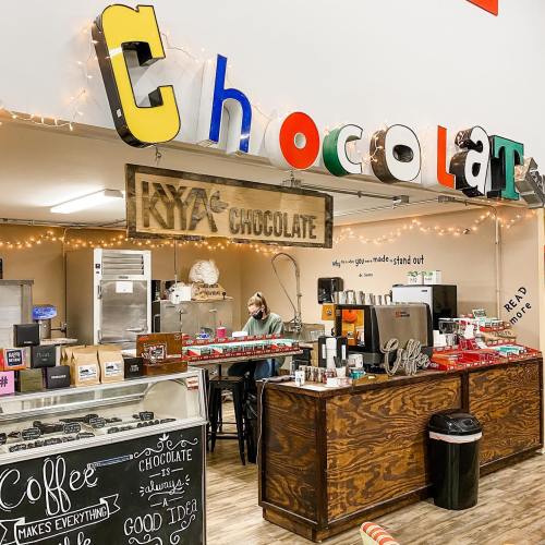 A cozy chocolate shop with colorful signage, a barista working, and shelves filled with chocolate products and coffee.
