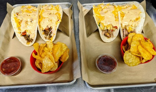 Two trays with tacos topped with cheese and sauce, served with a side of chips and salsa in red bowls.