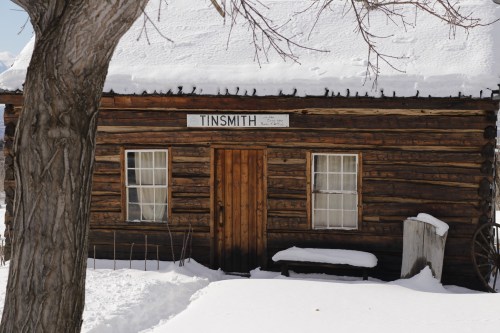 A rustic log cabin with a sign reading "TINSMITH," surrounded by snow and a tree in the foreground.