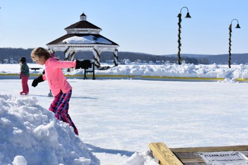 A girl in a pink sweater plays on a snowy ice rink near a gazebo, with another child in the background.