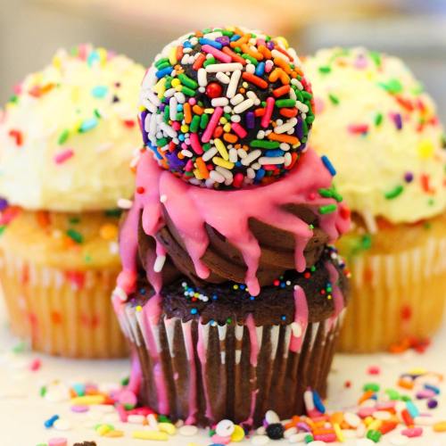 Three colorful cupcakes with vibrant frosting and sprinkles, featuring a large, sprinkles-covered cake ball on top.